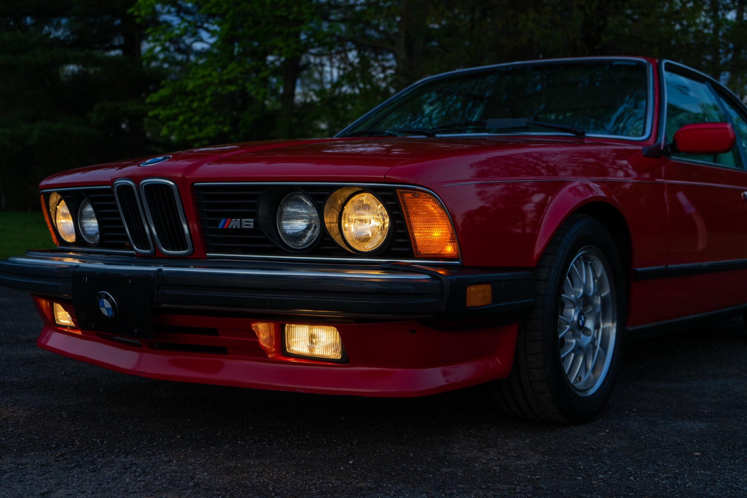 Front of a red BMW M6 at dusk with headlights and fog lamps lit