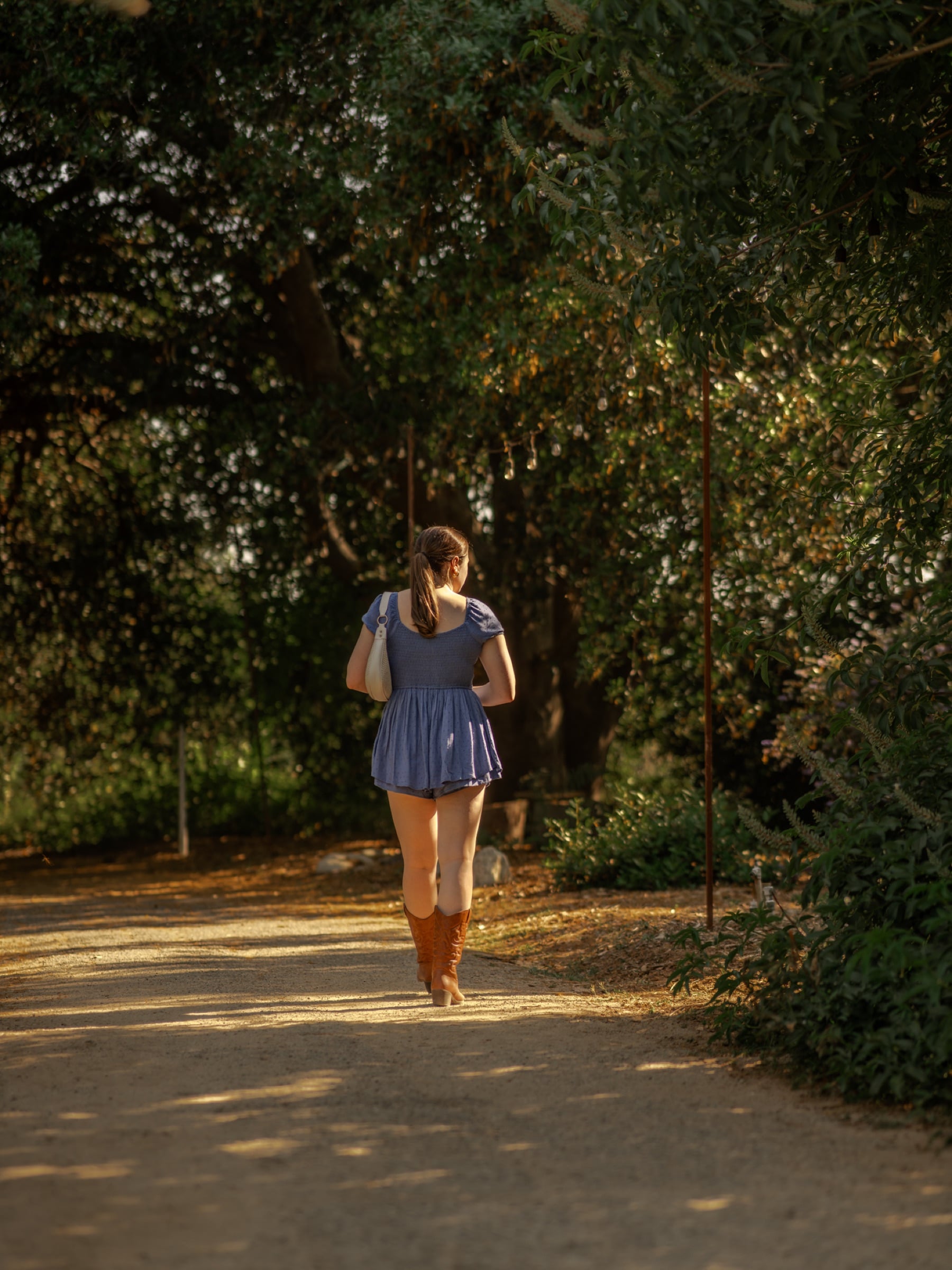 Woman in a blue dress and boots walking a sunlit tree-lined garden path