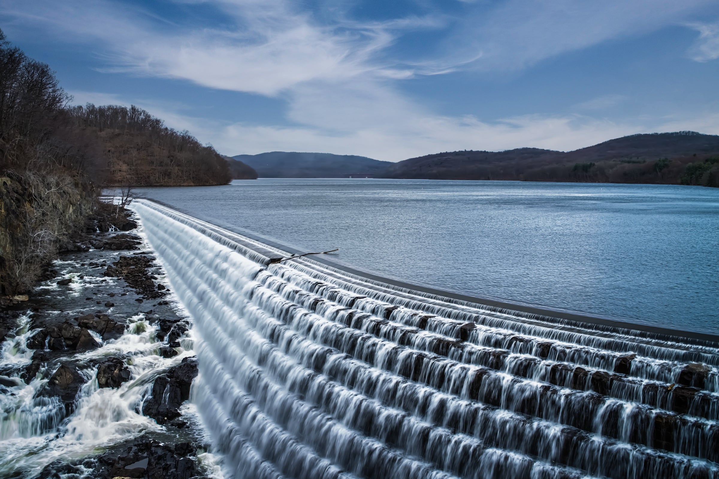 Wide stepped dam spillway flowing into a lake below forested hills