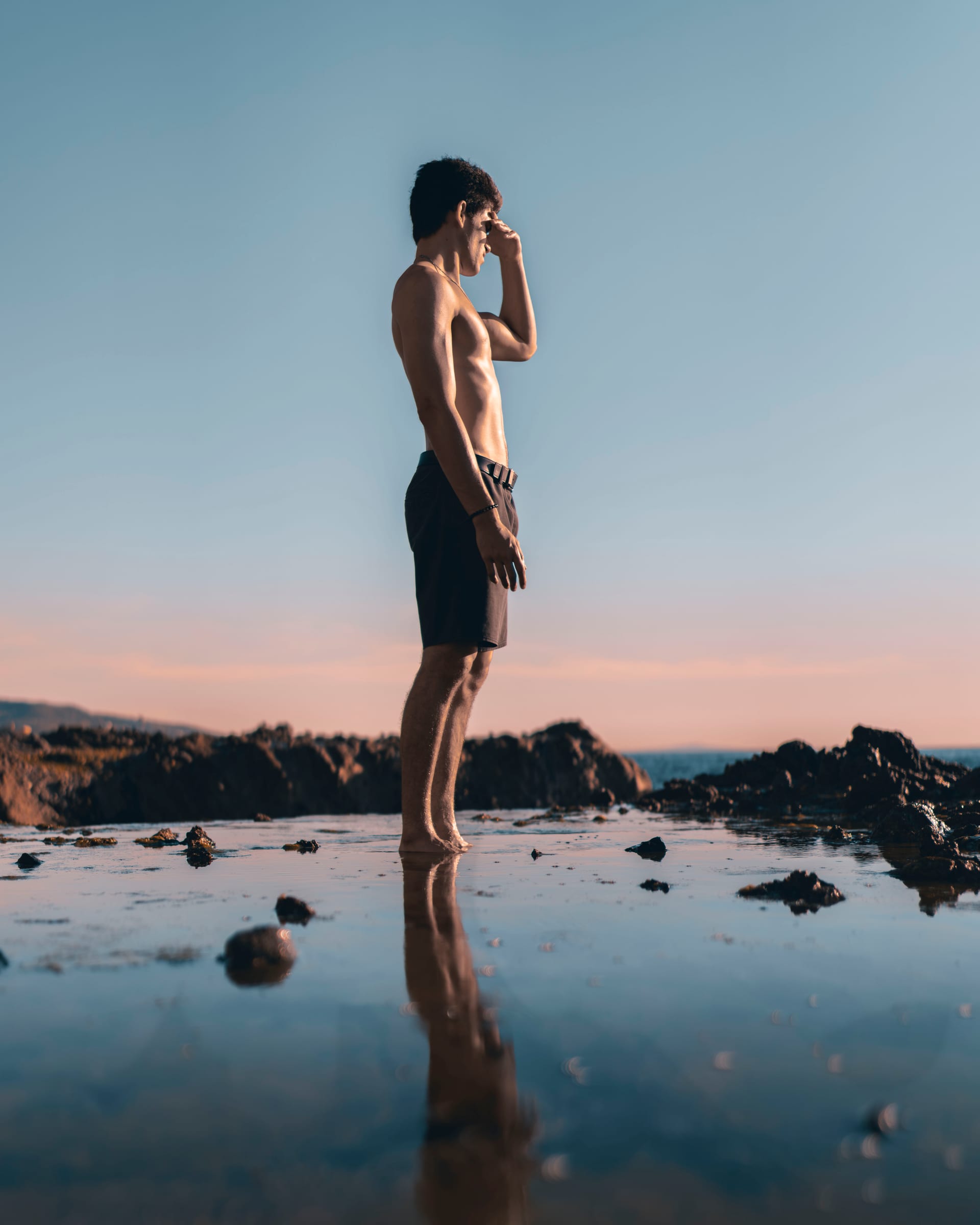 A young man standing barefoot on a glassy tide-pool flat at sunset, gazing toward the sea