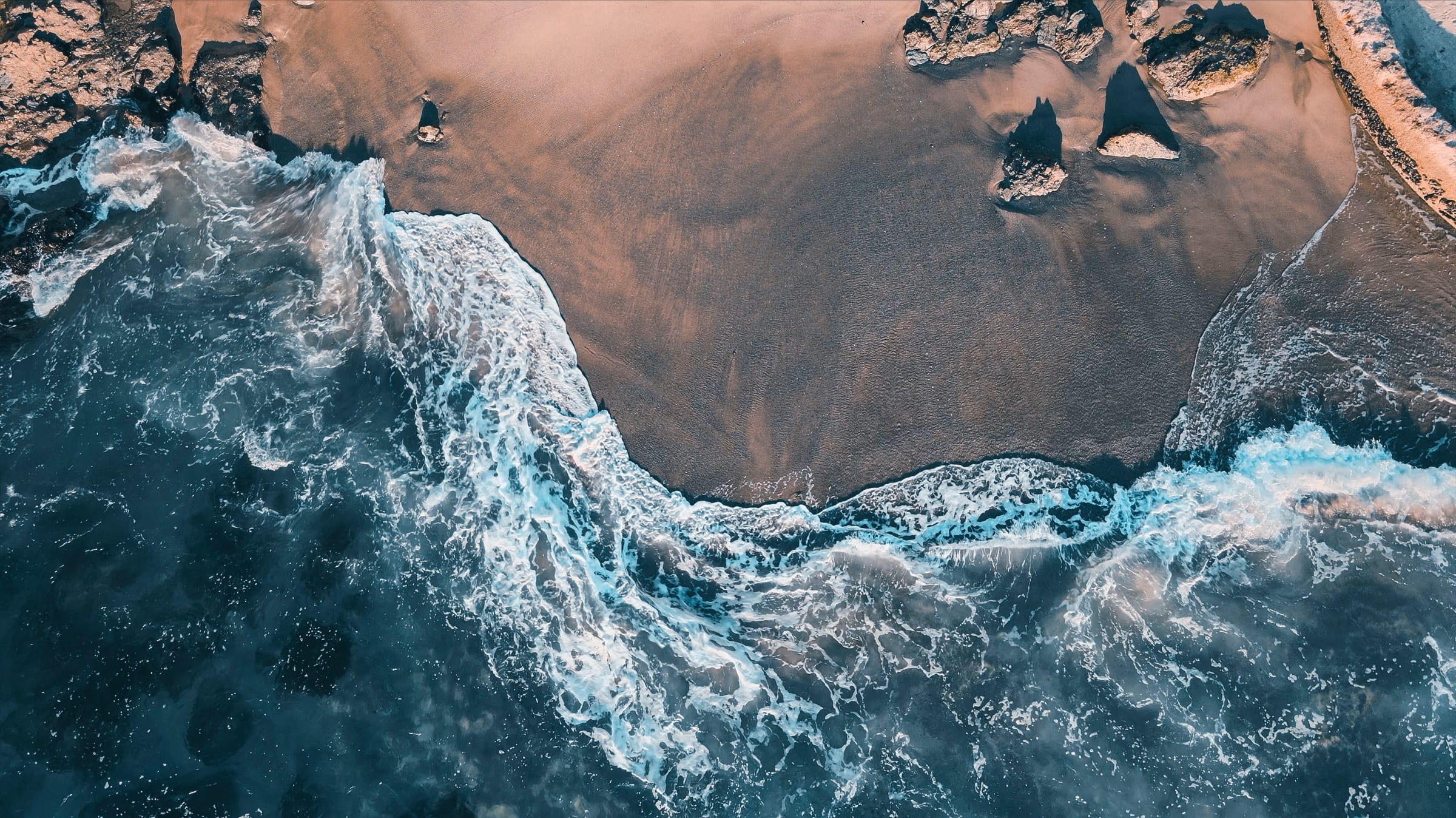 Wide aerial of foaming surf sweeping across a curved sandy shoreline