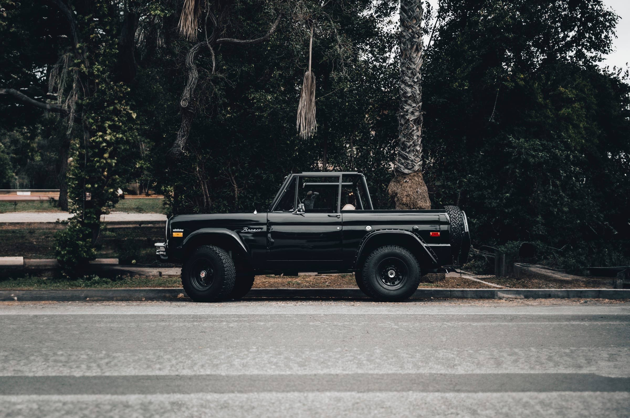 Side profile of a black vintage Ford Bronco parked beneath trees
